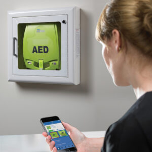 Eine Dame steht vor einem Defibrillator und hat ein Handy mit der Plus Trac App iin der Hand